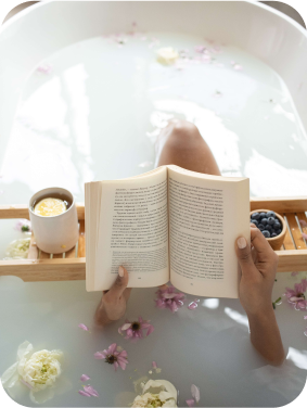 Mulher em uma banheira com pétalas de rosa, com apenas mãos e pernas aparentes, segurando um livro e com uma bandeija com uvas e chá.