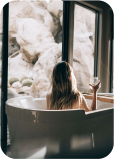 Busto de uma mulher de costas em uma banheira de espumas segurando uma taça e de frente a uma porta de vidro com grandes pedras atrás.